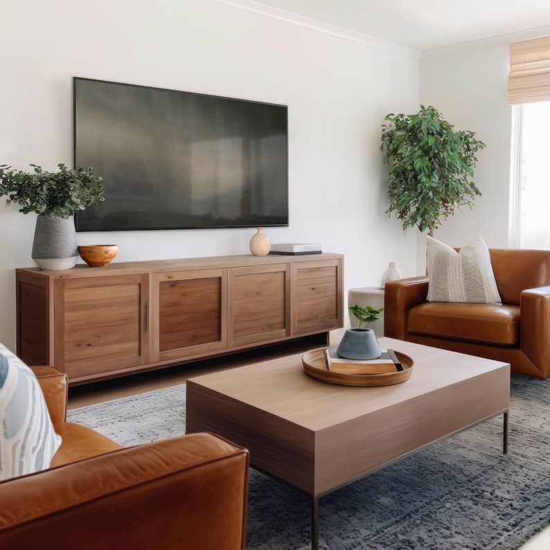 May include: A living room with a large flat screen TV mounted on the wall above a wooden entertainment center. There is a brown leather sofa with a throw pillow, a coffee table with a wooden tray and a ceramic vase, and a potted plant in the corner.