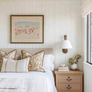 May include: A white bedroom with a bed, a nightstand, and a framed print of a beach scene. The bed has a white headboard and a white comforter. The nightstand is made of wood and has three drawers. The print is in a gold frame and shows a beach scene with people and umbrellas.