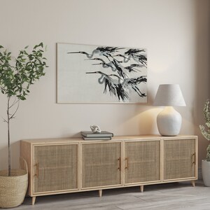 May include: A framed artwork depicts a flock of birds in flight, rendered in shades of black and gray against a light background. The artwork hangs above a light wood cabinet with woven door panels. A white lamp and potted plants complete the scene.