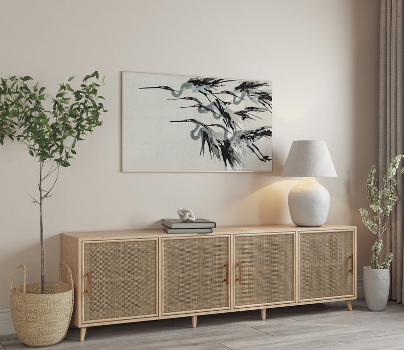 May include: A framed artwork depicts a flock of birds in flight, rendered in shades of black and gray against a light background. The artwork hangs above a light wood cabinet with woven door panels. A white lamp and potted plants complete the scene.