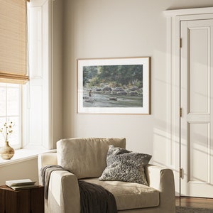 May include: A framed watercolor painting of a river scene with a person fishing in the water. The painting is in a light wood frame and is hanging on a wall in a room with a beige armchair and a window with a bamboo blind.