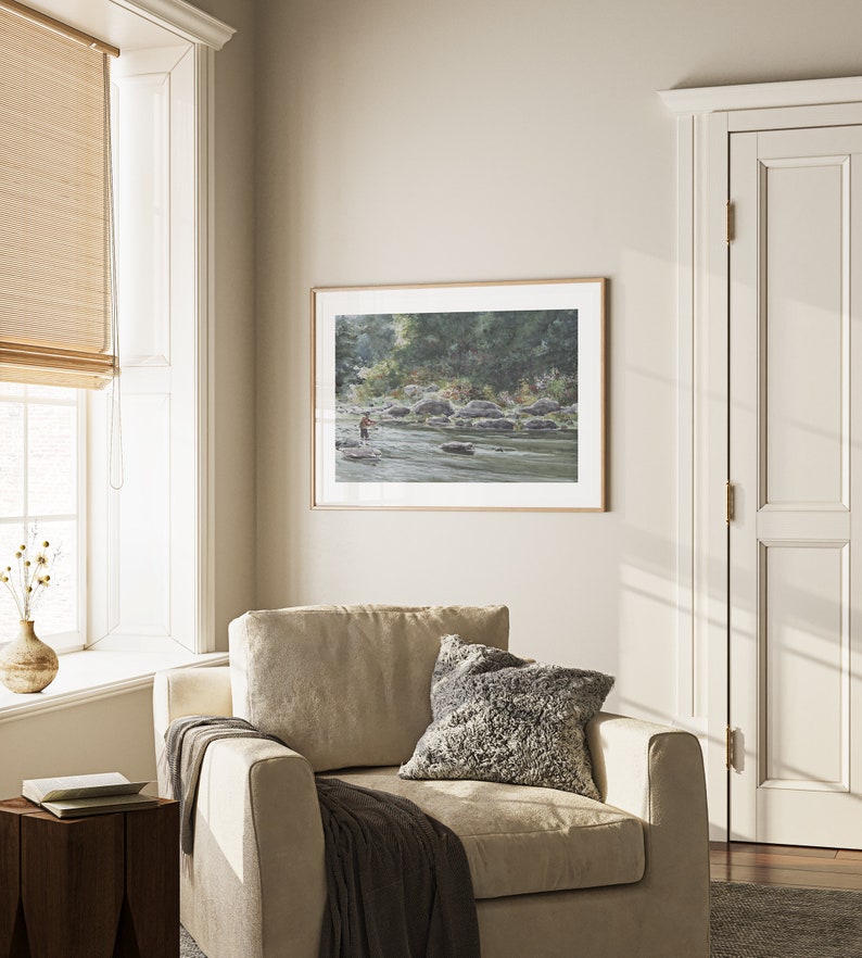 May include: A framed watercolor painting of a river scene with a person fishing in the water. The painting is in a light wood frame and is hanging on a wall in a room with a beige armchair and a window with a bamboo blind.