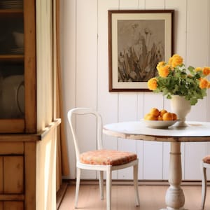 May include: A white round table with a white pedestal base, two white chairs, and a bouquet of yellow flowers in a white vase. The table is set with a white bowl of oranges.