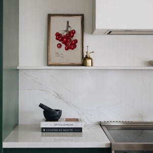 May include: A white kitchen countertop with a framed botanical print of red cherries, a gold metal spray bottle, a black mortar and pestle, and three cookbooks stacked on top of each other. The cookbooks are titled "Ottolenghi", "CUISINER EN BEAUT&Eacute;", and "CUISINER EN BEAUT&Eacute;".