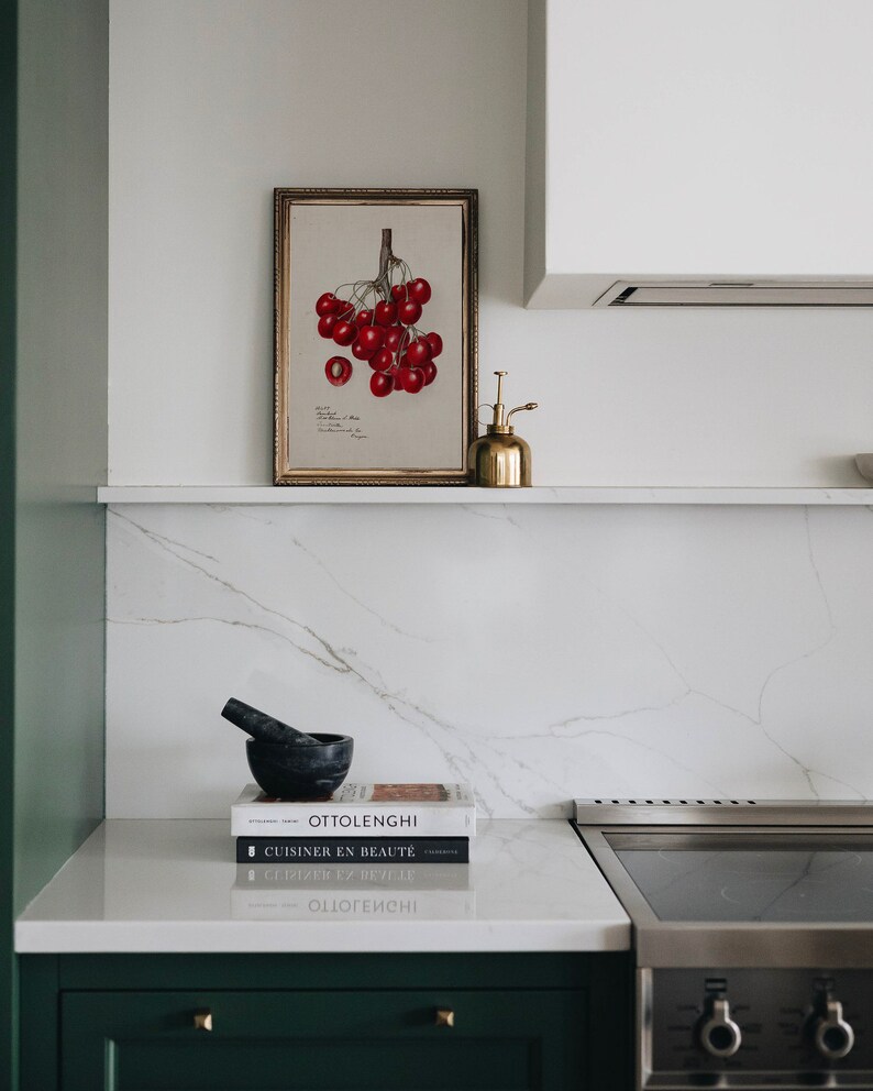 May include: A white kitchen countertop with a framed botanical print of red cherries, a gold metal spray bottle, a black mortar and pestle, and three cookbooks stacked on top of each other. The cookbooks are titled "Ottolenghi", "CUISINER EN BEAUT&Eacute;", and "CUISINER EN BEAUT&Eacute;".