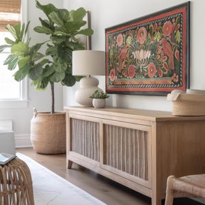Puede incluir: Una consola de madera clara con dos puertas de rat&aacute;n. La consola est&aacute; decorada con una gran pintura enmarcada de flores y p&aacute;jaros coloridos. Una gran planta de ficus lyrata en maceta se encuentra a la izquierda de la consola.