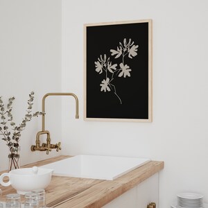 May include: A framed black and white print of a stylized floral design. The print is hanging on a white wall above a white sink with a gold faucet.