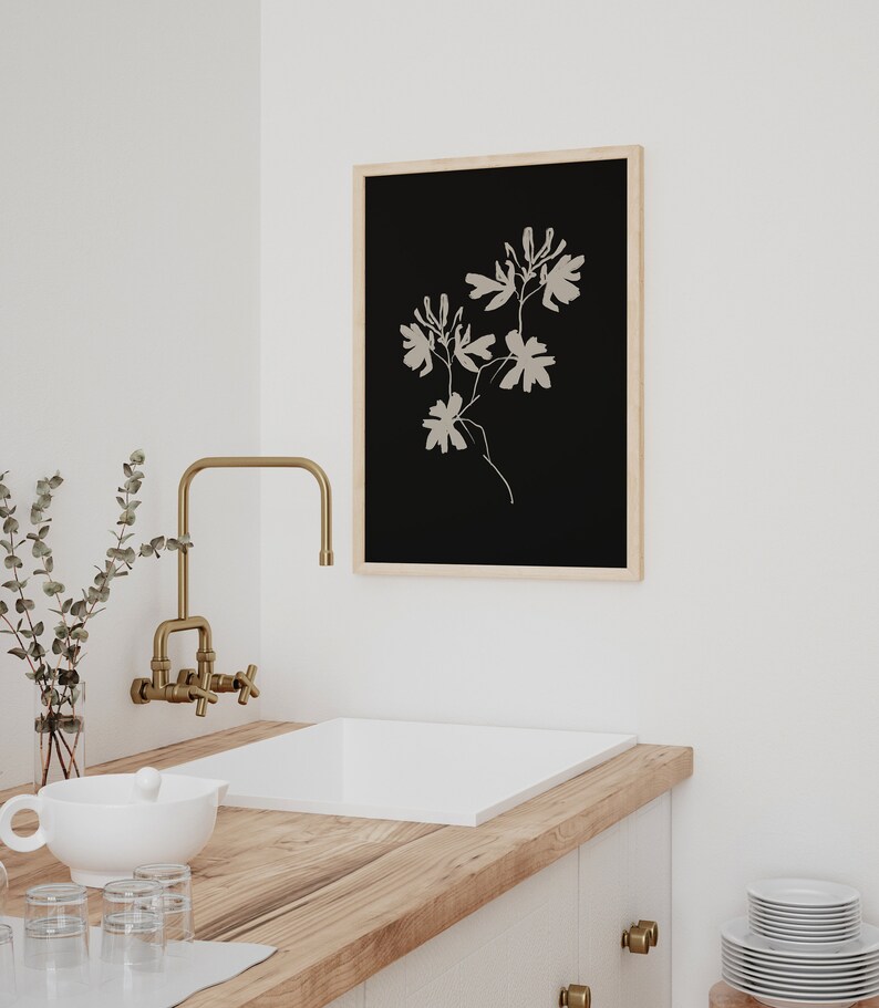 May include: A framed black and white print of a stylized floral design. The print is hanging on a white wall above a white sink with a gold faucet.