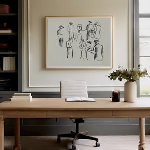 Op de afbeelding: Een licht houten bureau met een ingelijste schets van figuren erboven. Een witte vaas met groen en een kaars staan op het bureau. Een grijze boekenkast staat links en een raam rechts.