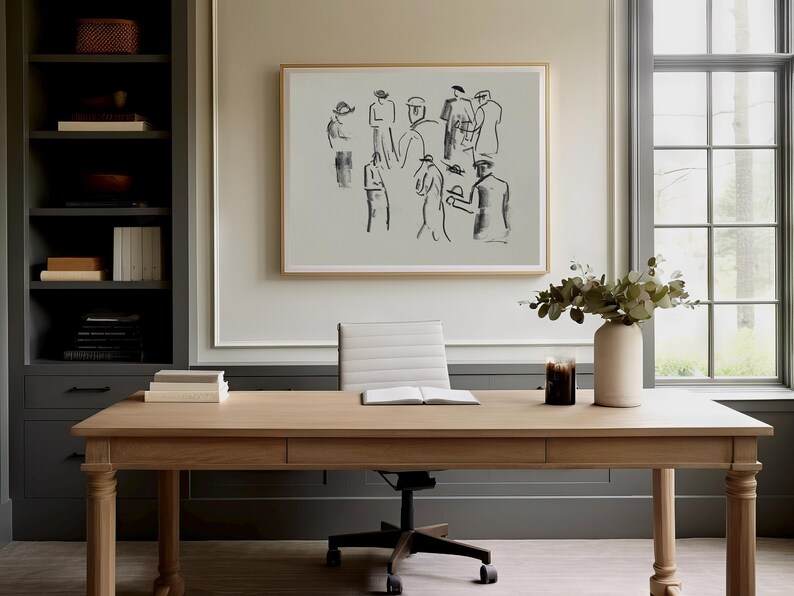 Op de afbeelding: Een licht houten bureau met een ingelijste schets van figuren erboven. Een witte vaas met groen en een kaars staan op het bureau. Een grijze boekenkast staat links en een raam rechts.