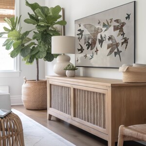 May include: A light wood cabinet with woven panels, a lamp, and a framed artwork featuring bird silhouettes. A large potted plant in a woven basket and a small succulent in a pot are also visible. The room has a neutral color palette.