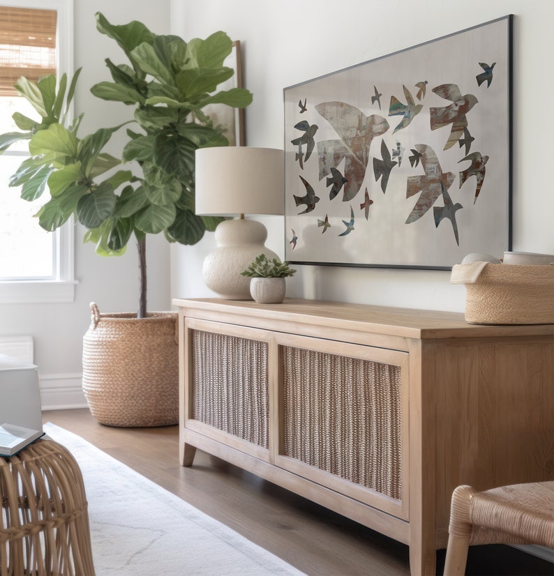 May include: A light wood cabinet with woven panels, a lamp, and a framed artwork featuring bird silhouettes. A large potted plant in a woven basket and a small succulent in a pot are also visible. The room has a neutral color palette.