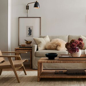 May include: A living room scene featuring a light-colored sofa with decorative pillows, a woven coffee table, and a wooden side chair. A framed artwork and a brass lamp are mounted on the wall. A vase of dried flowers adds a touch of color.