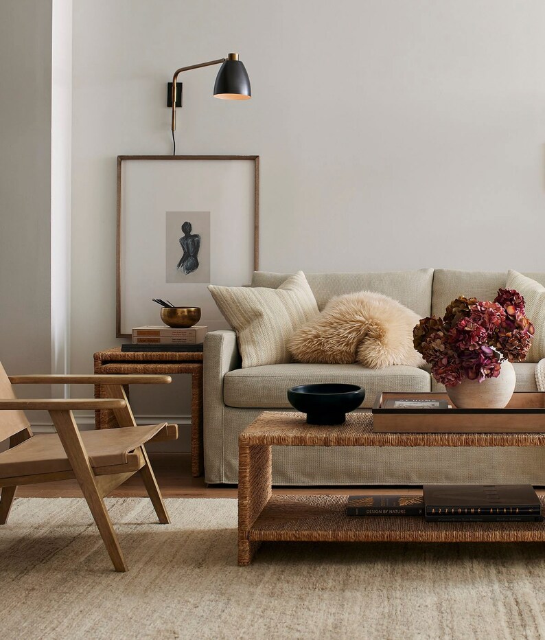 May include: A living room scene featuring a light-colored sofa with decorative pillows, a woven coffee table, and a wooden side chair. A framed artwork and a brass lamp are mounted on the wall. A vase of dried flowers adds a touch of color.