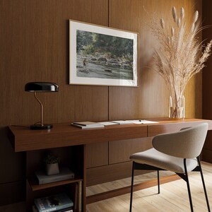 May include: A wooden desk with a black lamp, a chair, and a framed watercolor painting of a river scene. There are books on a shelf below the desk.