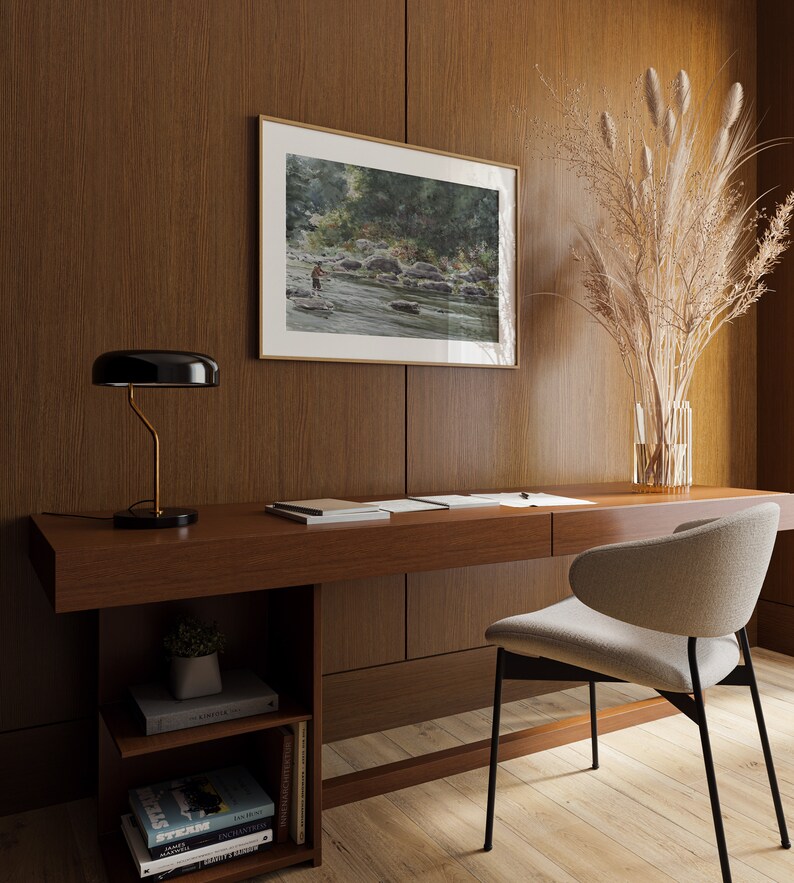 May include: A wooden desk with a black lamp, a chair, and a framed watercolor painting of a river scene. There are books on a shelf below the desk.