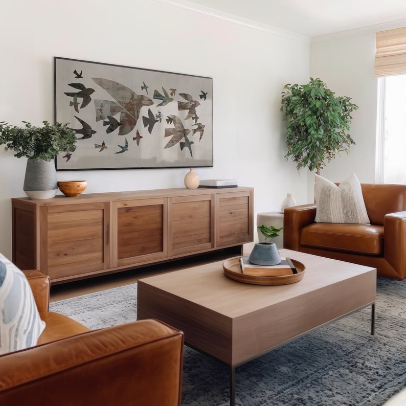 May include: A living room scene featuring a large wooden cabinet, a coffee table, and two leather armchairs. A framed artwork of birds in flight hangs above the cabinet. Decorative vases and a plant add to the room's aesthetic.