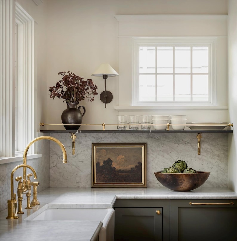 May include: A kitchen with white walls and marble countertops. A gold faucet is mounted on the counter next to a white sink. There is a shelf above the counter with a framed picture, glasses, and a bowl of artichokes. The cabinets are dark green with gold hardware.