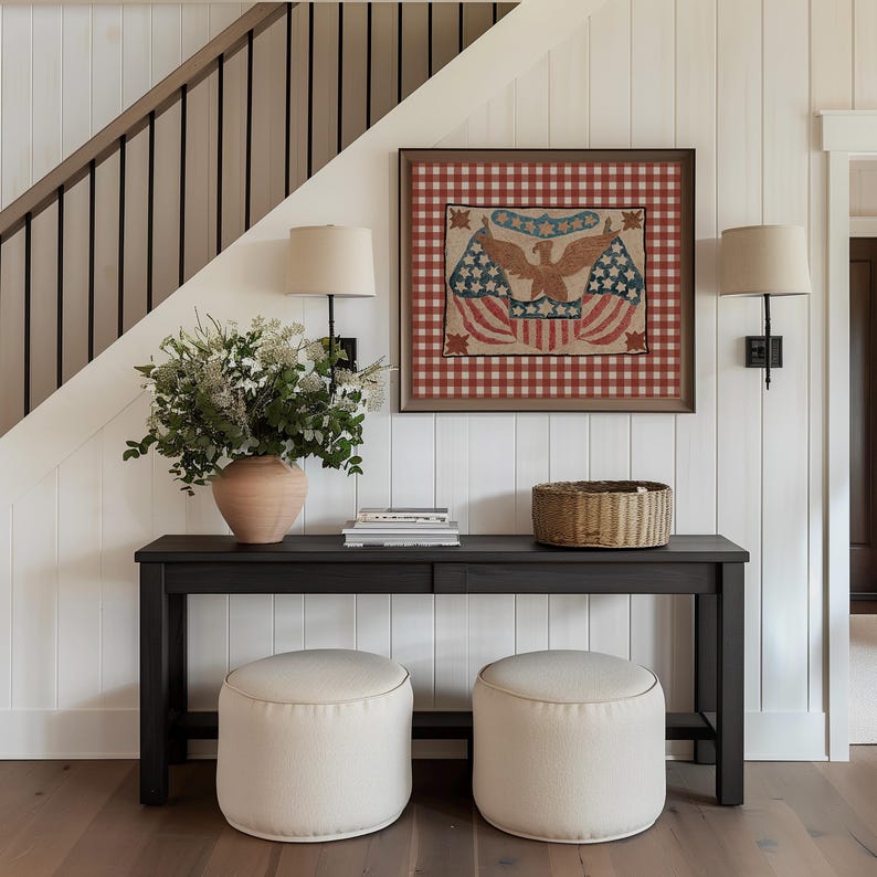Puede incluir: Un cuadro patri&oacute;tico enmarcado con un dise&ntilde;o de &aacute;guila y bandera, sobre un fondo a cuadros rojos y blancos. La habitaci&oacute;n cuenta con una consola gris oscura, dos otomanas color crema y un jarr&oacute;n con flores. Dos apliques de pared con pantallas beige flanquean la obra.