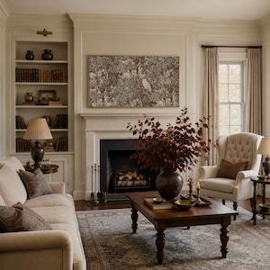 May include: A living room featuring a beige sofa with brown pillows, a dark wood coffee table with a large vase of red leaves, a wingback chair, a fireplace with a framed owl print above it, and built-in bookshelves filled with books.