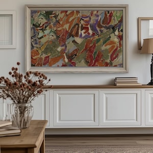 May include: Abstract painting in a light wooden frame. The artwork uses orange, red, green, and purple. It hangs above a white cabinet with a wooden top. A vase of dried flowers sits on a wooden table in a room with a patterned rug.