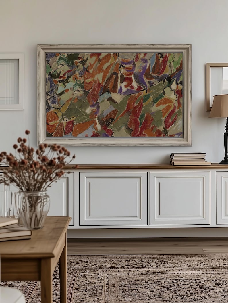 May include: Abstract painting in a light wooden frame. The artwork uses orange, red, green, and purple. It hangs above a white cabinet with a wooden top. A vase of dried flowers sits on a wooden table in a room with a patterned rug.