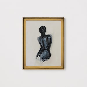 May include: Abstract art print featuring a figure's back in shades of black and gray. The artwork is framed in a gold-colored, ornate frame. The background is a neutral beige color, creating a contrast with the dark figure.