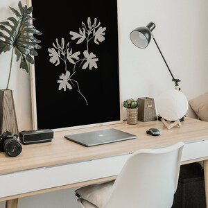May include: A minimalist black and white framed print of a stylized floral design hangs above a wooden desk with a laptop, a lamp, and a white chair.