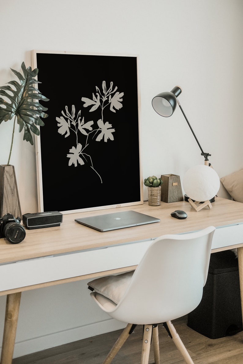 May include: A minimalist black and white framed print of a stylized floral design hangs above a wooden desk with a laptop, a lamp, and a white chair.