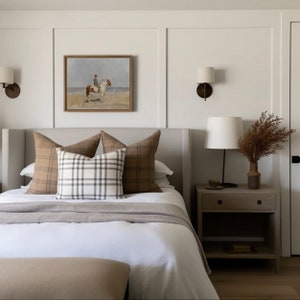 May include: A white bed with a gray headboard and a white duvet cover. There are three pillows on the bed, two with a brown and white plaid pattern and one with a white and gray plaid pattern. A brown and white painting hangs above the bed. A nightstand with a lamp and a vase of dried flowers sits next to the bed.