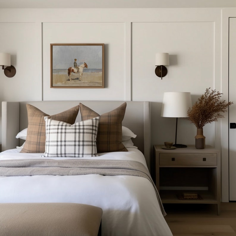 May include: A white bed with a gray headboard and a white duvet cover. There are three pillows on the bed, two with a brown and white plaid pattern and one with a white and gray plaid pattern. A brown and white painting hangs above the bed. A nightstand with a lamp and a vase of dried flowers sits next to the bed.