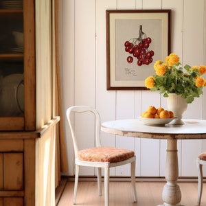 May include: A white round table with two white chairs sits in a room with white walls and wood floors. A vase of yellow flowers sits on the table, and a bowl of oranges sits next to it. A framed botanical print of red cherries hangs on the wall.