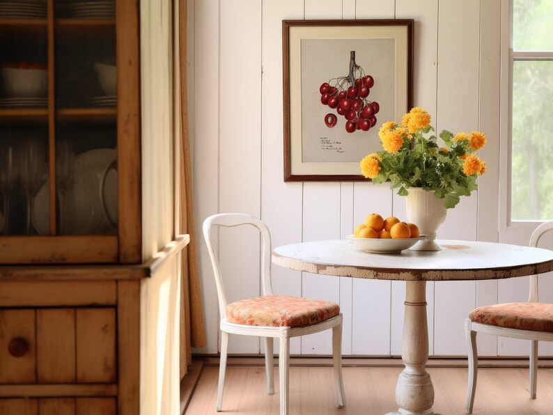 May include: A white round table with two white chairs sits in a room with white walls and wood floors. A vase of yellow flowers sits on the table, and a bowl of oranges sits next to it. A framed botanical print of red cherries hangs on the wall.