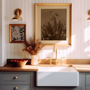May include: A white farmhouse kitchen sink with a gold tap and a wooden worktop. A framed print of a floral still life hangs above the sink.