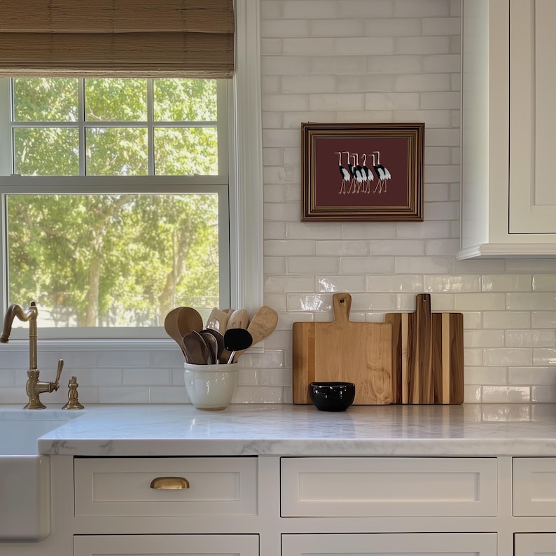 May include: A white kitchen with white subway tile backsplash, a window with a view of trees, and a framed print of birds on a red background. There are wooden cutting boards and a bowl on the counter.