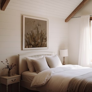 May include: A white bed with a white duvet and a beige throw blanket. The bed is in a bedroom with white walls and a large window with white curtains. There is a framed painting of flowers on the wall behind the bed.