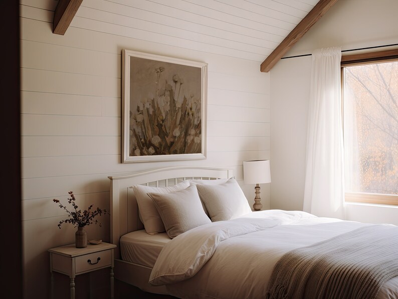 May include: A white bed with a white duvet and a beige throw blanket. The bed is in a bedroom with white walls and a large window with white curtains. There is a framed painting of flowers on the wall behind the bed.