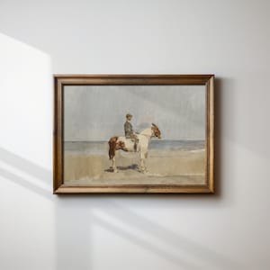 May include: A framed painting of a person riding a brown and white horse on a beach. The person is wearing a dark jacket and a hat. The painting is in a wooden frame.