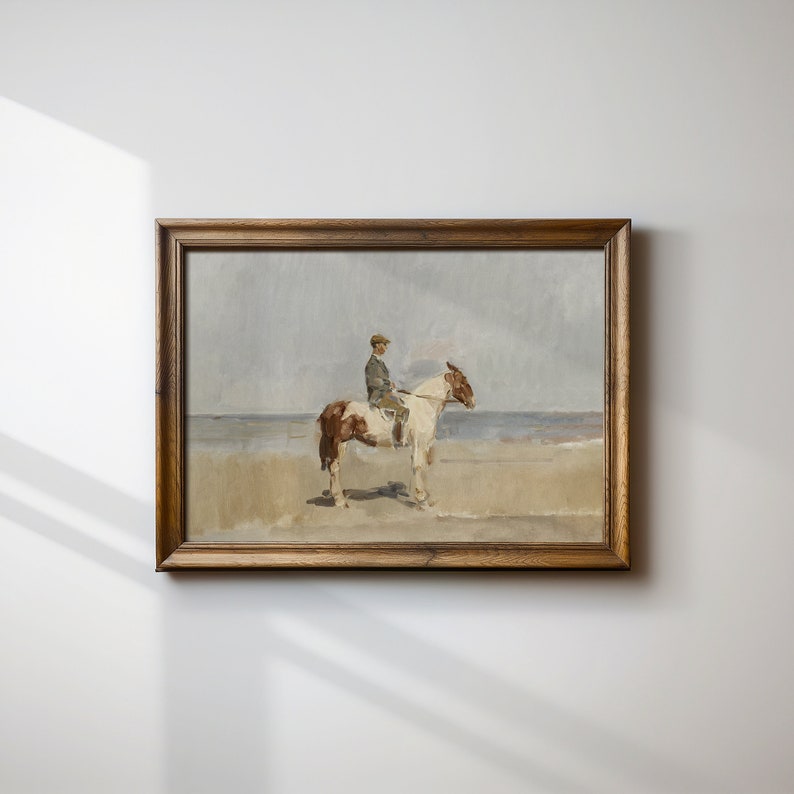 May include: A framed painting of a person riding a brown and white horse on a beach. The person is wearing a dark jacket and a hat. The painting is in a wooden frame.