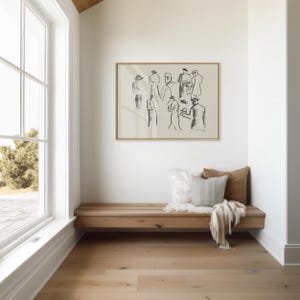 Op de afbeelding: Een ingelijst kunstwerk met een schets van mensen hangt boven een houten bank met kussens en een deken. De kamer heeft een groot raam en een lichtgekleurde houten vloer.