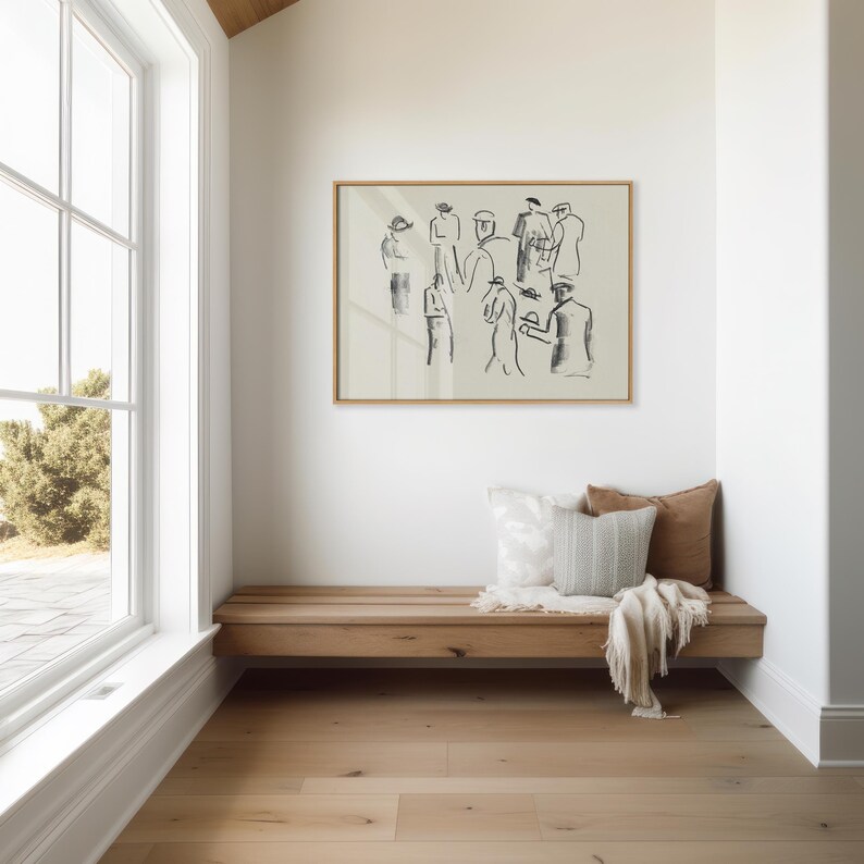 Op de afbeelding: Een ingelijst kunstwerk met een schets van mensen hangt boven een houten bank met kussens en een deken. De kamer heeft een groot raam en een lichtgekleurde houten vloer.