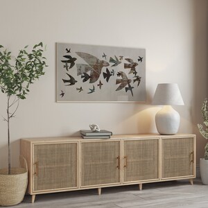 May include: A framed artwork featuring a flock of birds in flight, rendered in muted tones of brown, gray, and blue. The artwork hangs above a light wood cabinet with woven cane door panels and brass handles. A white lamp and potted plants complete the scene.