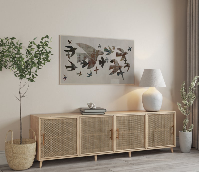 May include: A framed artwork featuring a flock of birds in flight, rendered in muted tones of brown, gray, and blue. The artwork hangs above a light wood cabinet with woven cane door panels and brass handles. A white lamp and potted plants complete the scene.