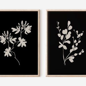 May include: Two framed prints with black backgrounds and white floral silhouettes. The print on the left features a single stem with multiple flowers and leaves. The print on the right features a single stem with multiple leaves and buds.