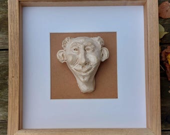 May include: A framed ceramic sculpture of a person's face with a whimsical expression. The sculpture is painted in a light brown colour and is mounted on a brown background.