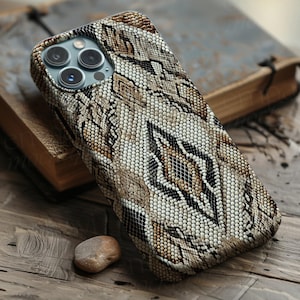 Puede incluir: Funda de teléfono con estampado de piel de serpiente en tonos marrón, beige y negro. Descansa sobre una superficie de madera junto a una pequeña piedra y un libro abierto con páginas envejecidas. La funda tiene un aspecto texturizado.