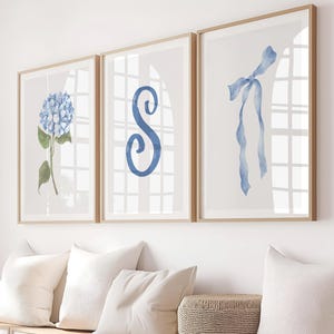 Pode incluir: Três impressões de arte emolduradas com designs em aquarela. As impressões apresentam uma hortênsia azul, uma letra "S" azul e uma fita azul. As molduras são de cor castanho claro e o fundo é um branco sujo neutro.