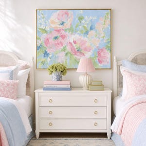 May include: A rectangular wall art piece featuring an impressionistic floral painting with soft pink, blue, and green brushstrokes. The artwork is framed in a thin gold border and hangs above a white three-drawer dresser with round gold knobs.