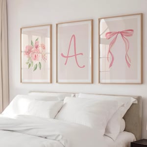 Könnte beinhalten: Drei gerahmte Kunstdrucke für die Wand. Einer zeigt einen rosa Blumenstrauß, ein weiterer den rosa Buchstaben "A" und der dritte eine rosa Schleife. Die Drucke werden über einem Bett mit weißer Bettwäsche ausgestellt. Die Rahmen sind hellbraun.