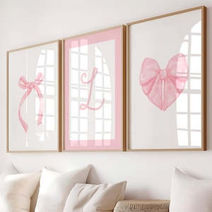 May include: Three framed prints with pink watercolor illustrations. The left print features a pink bow, the middle print features the letter L, and the right print features a pink bow in the shape of a heart.